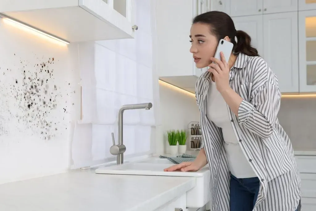 Woman on phone in a bright kitchen, leaning over a white counter near a sink.