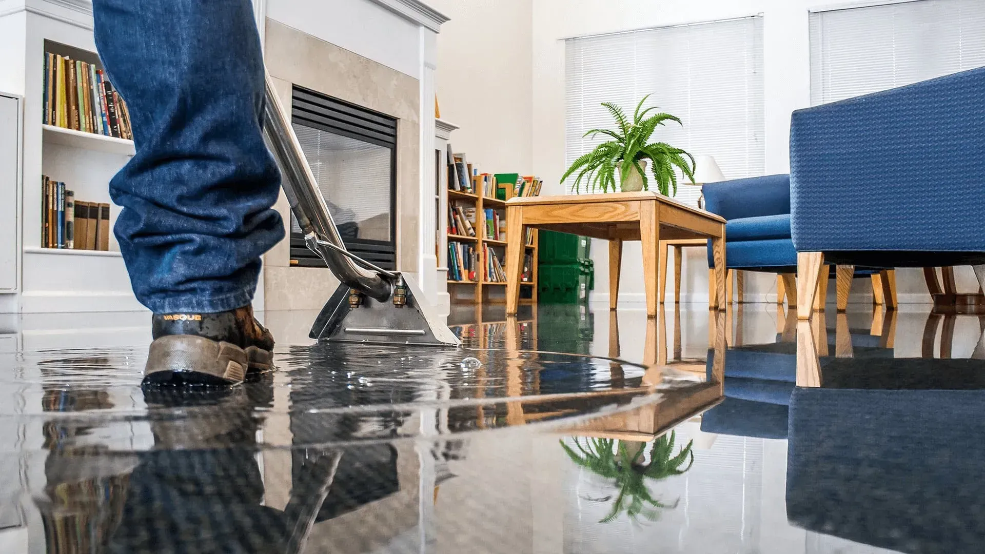 Person vacuuming a shiny living room floor near blue chairs and a wooden table with plants