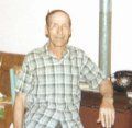 An individual wearing a green and white plaid short-sleeved shirt sits indoors near a metallic pole.