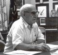 A person in a striped, short-sleeved collared shirt sits at a desk with papers, looking to the side in a formal setting.