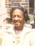 Smiling person wearing a light-colored jacket and a long necklace, standing outdoors in front of a stone building.
