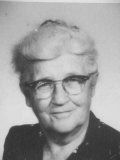 Black-and-white portrait of a person with short, light hair wearing glasses and a dark top, looking toward the camera.