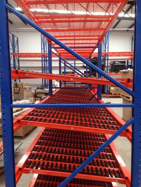 Estanterías de almacén con estructura azul y cintas transportadoras de rodillos rojos en un pasillo de almacenamiento industrial.