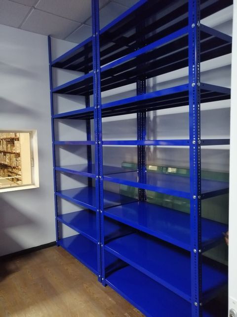 Estanterías metálicas azules en una habitación casi vacía con suelo de madera y una pequeña ventana a la izquierda.
