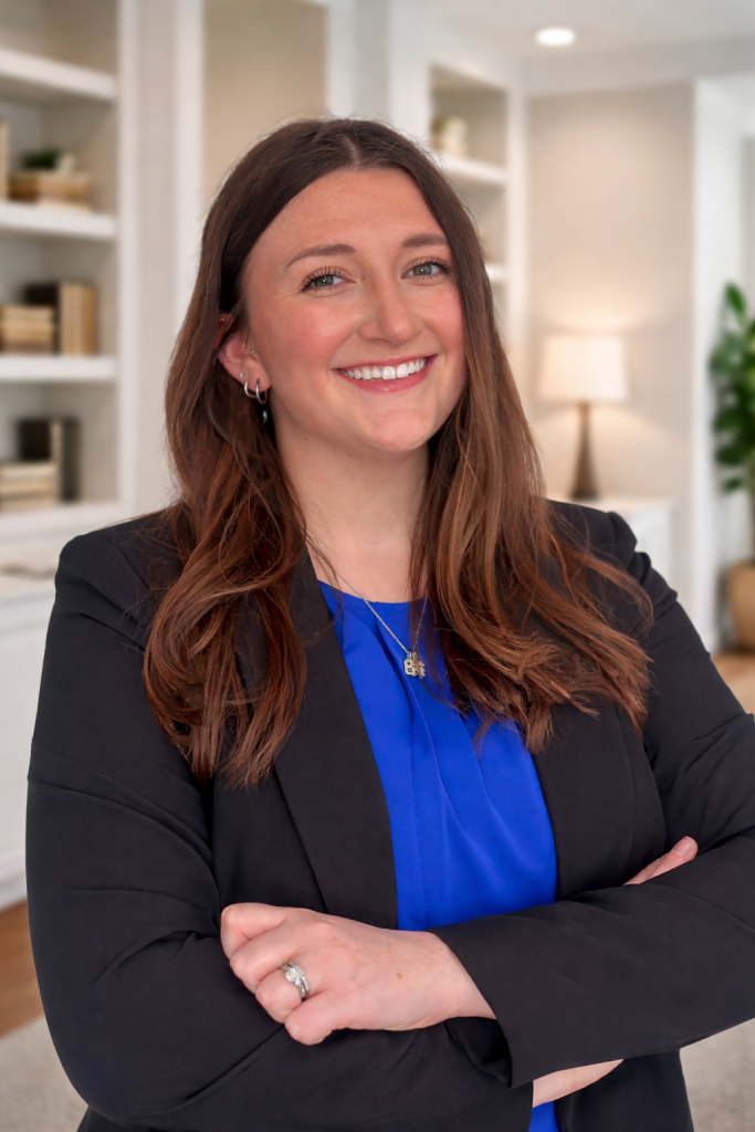 Smiling woman in a blue top and black blazer standing in a bright office with arms crossed