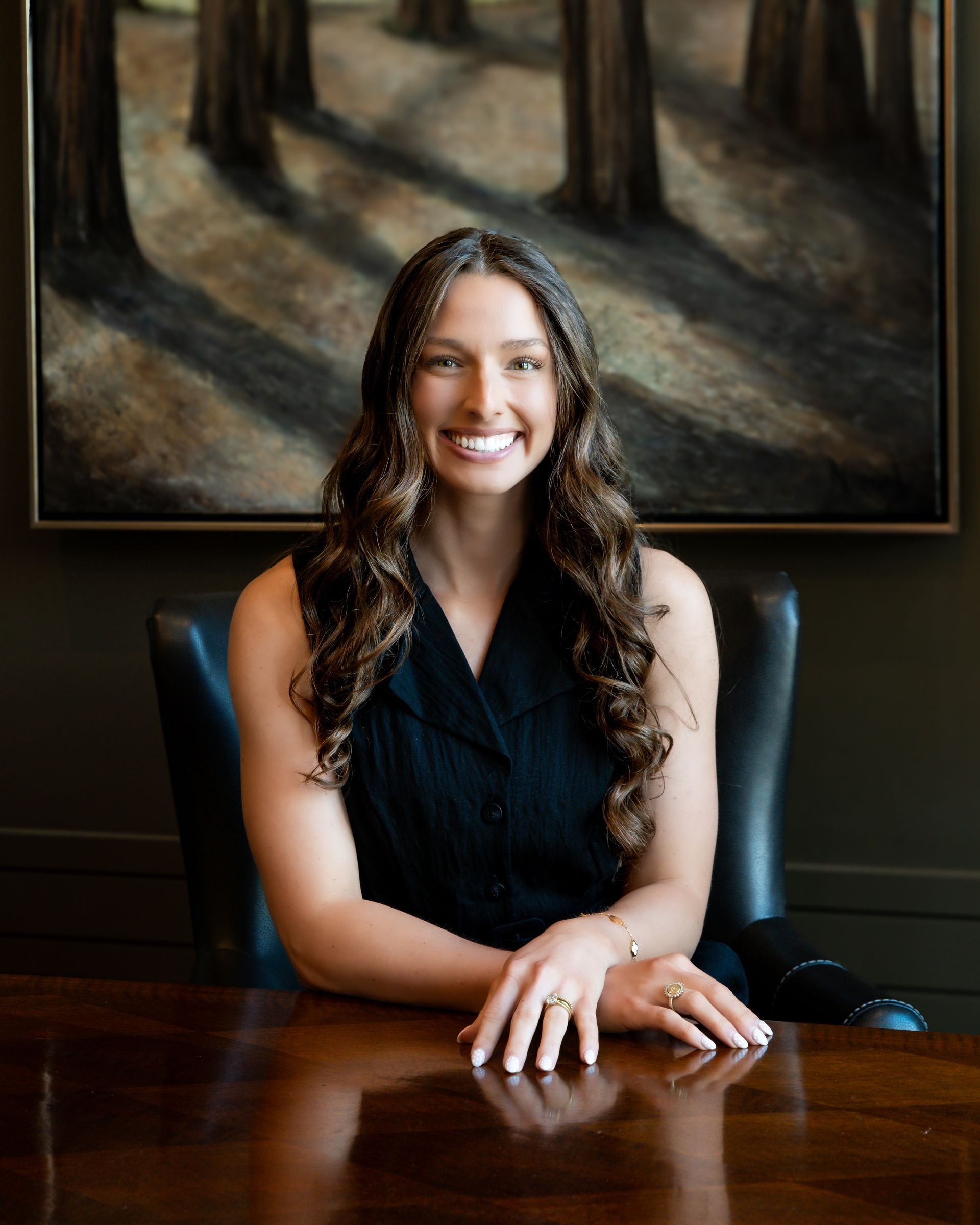 Woman in a black sleeveless top smiling at a wooden desk in a formal office setting.