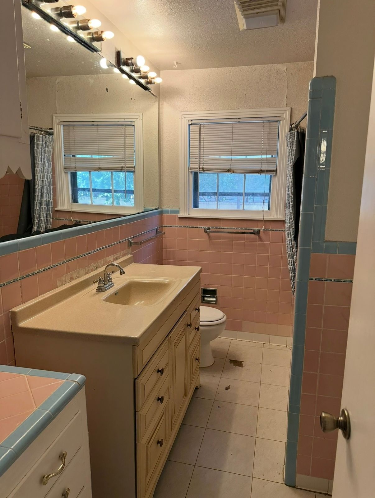 Small bathroom with pink tile, vanity, mirror, and two windows above the toilet.