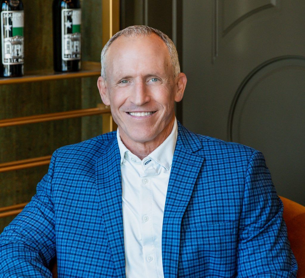 Smiling man in a blue checkered blazer seated indoors near shelves with bottles.