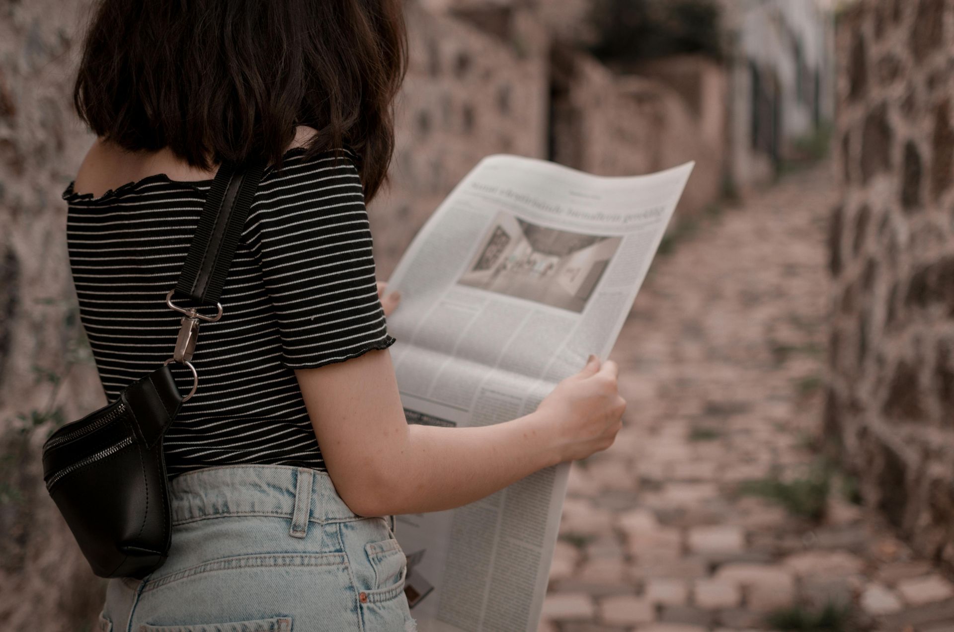 A person in a striped shirt and jeans reads a newspaper while walking down a narrow stone path.