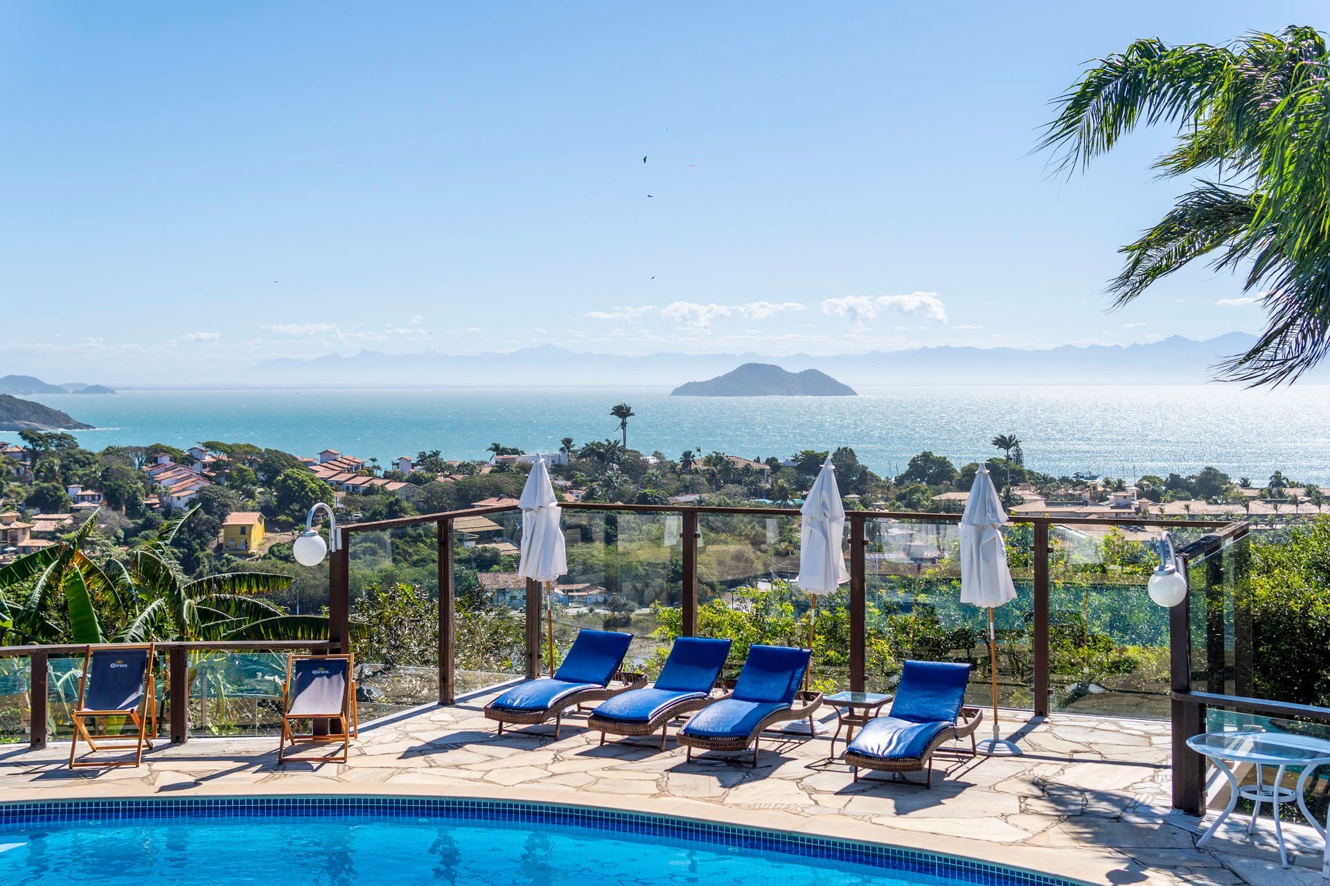 Espreguiçadeiras à beira da piscina com vista para o oceano ensolarado e para a ilha, a partir de um resort na encosta de uma colina.