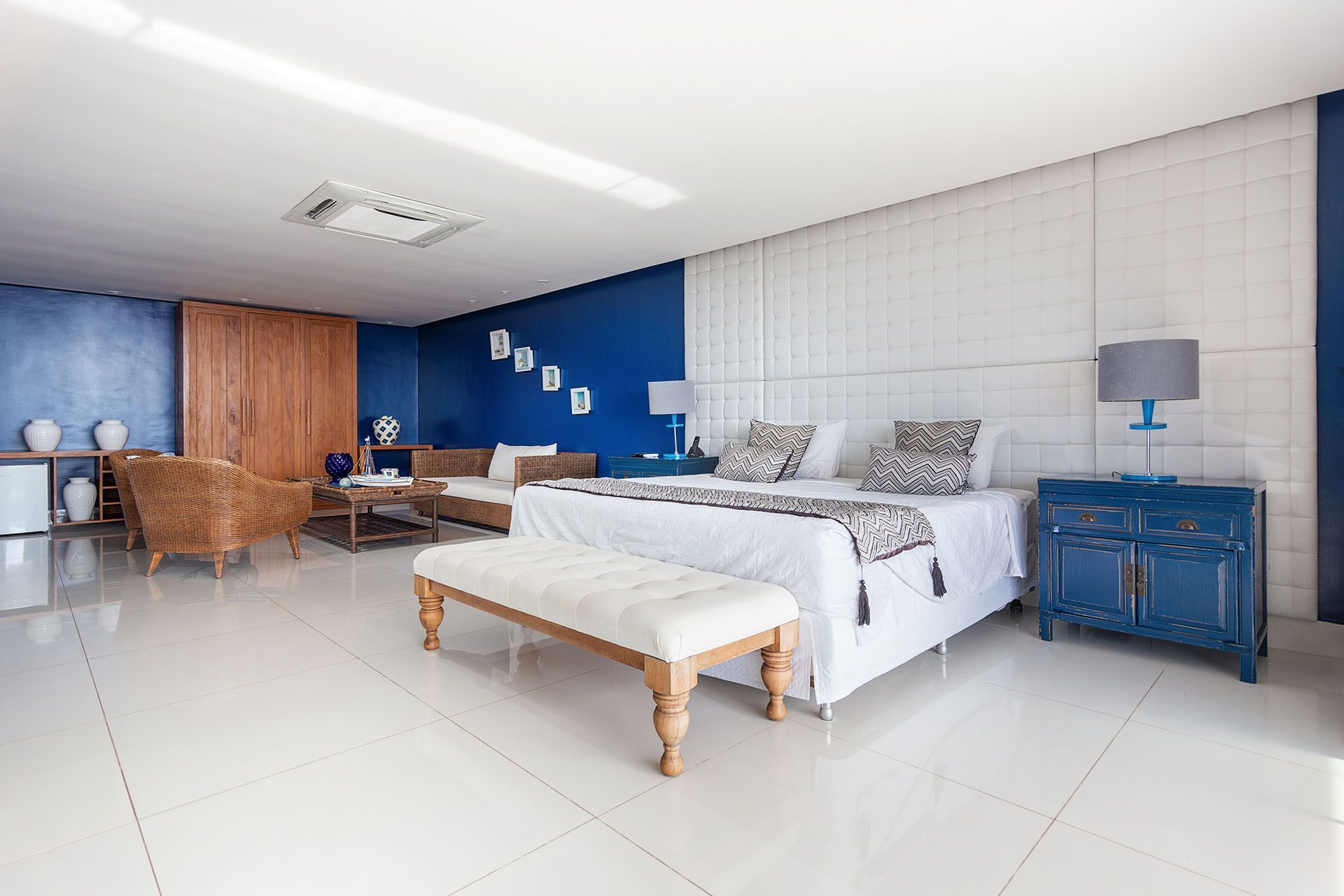 Quarto luminoso com cama branca, paredes com detalhes em azul, cadeira de vime e mesa de cabeceira azul.