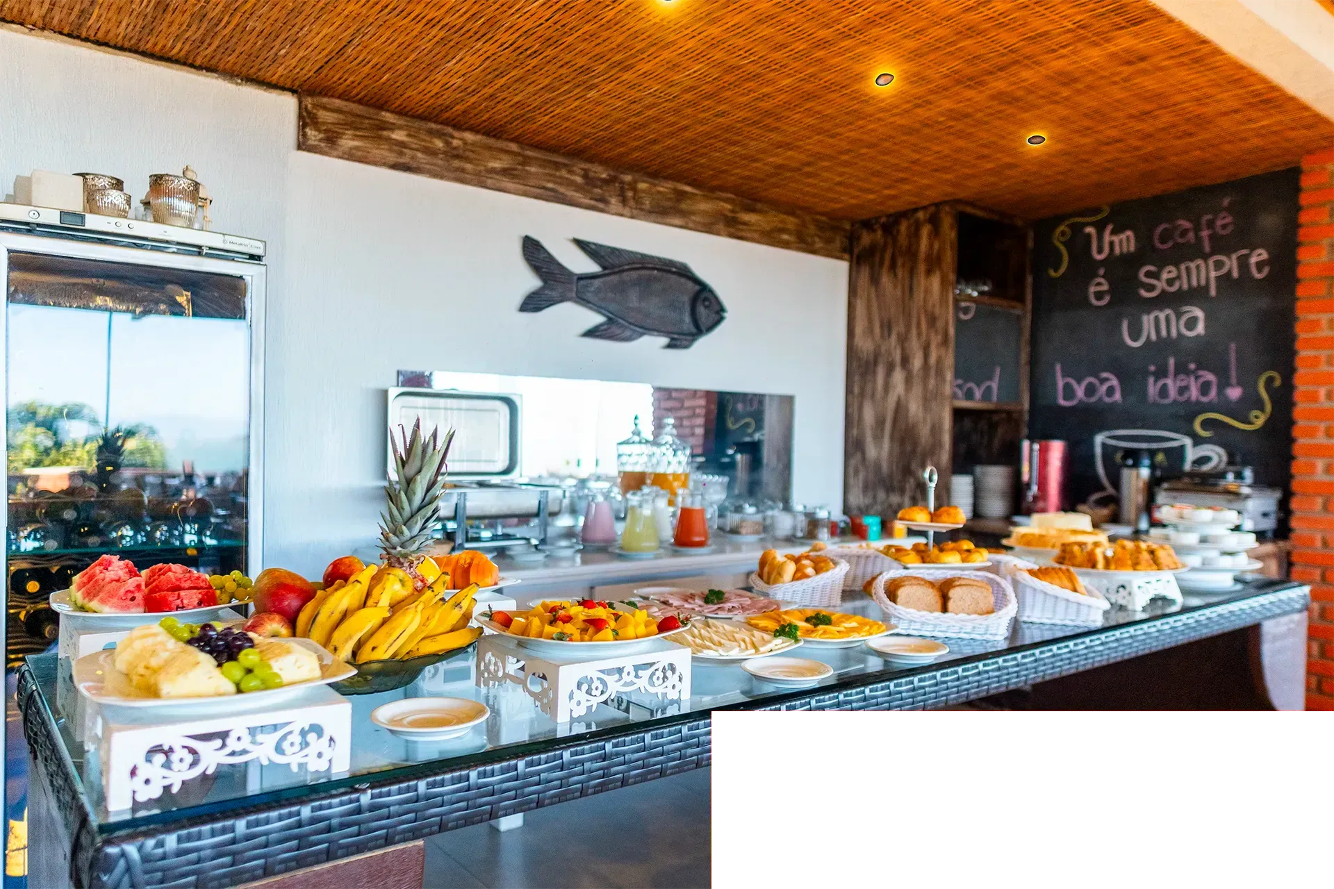 Mesa de buffet com doces, frutas e bebidas em um café iluminado; cardápio escrito em lousa na parede.