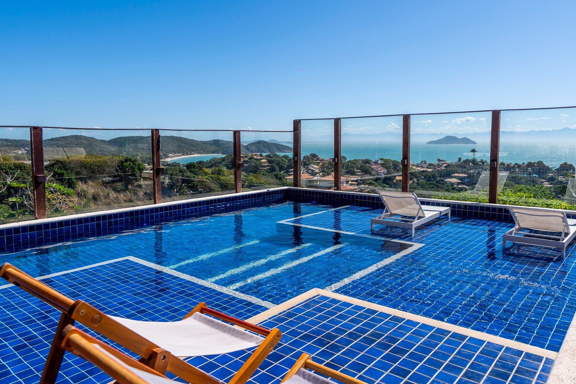 Piscina na cobertura com azulejos azuis, espreguiçadeiras e vista para o mar além do guarda-corpo de vidro.