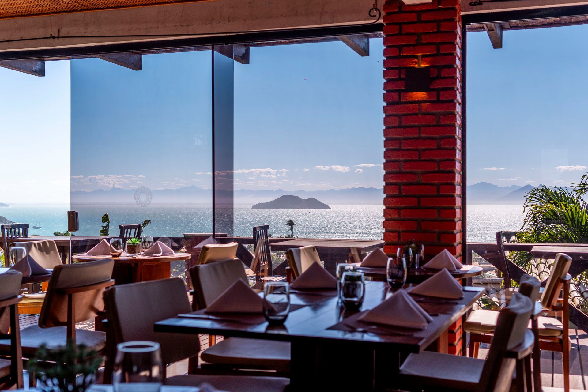 Pátio de restaurante à beira-mar com mesas postas, colunas de tijolos e vista para o oceano ao pôr do sol.