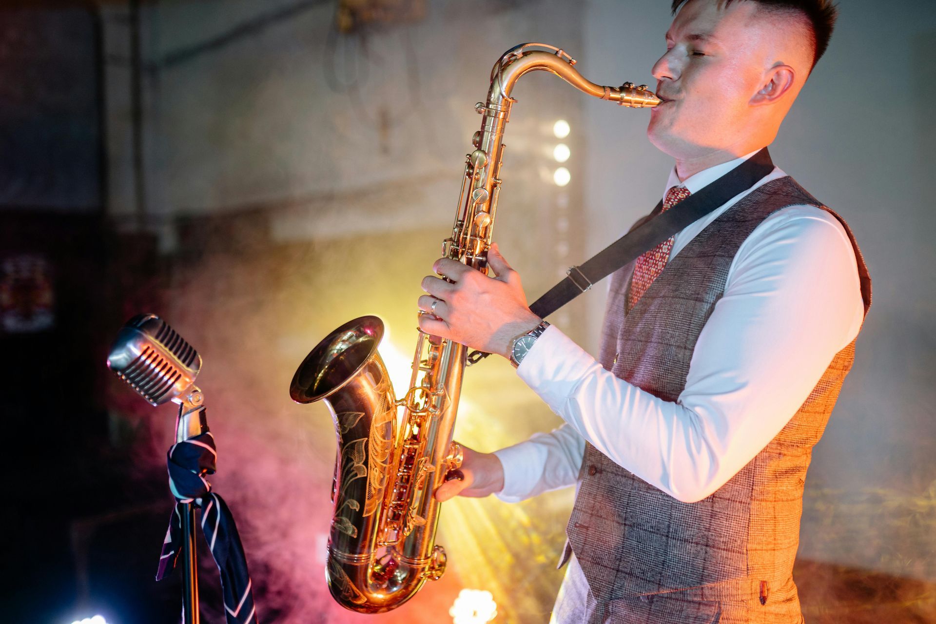 Homem de colete tocando saxofone com um microfone retrô; palco esfumaçado com luzes coloridas.