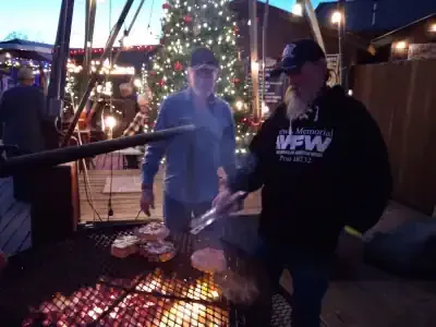 People grilling burgers at an outdoor holiday market beside a lit Christmas tree.