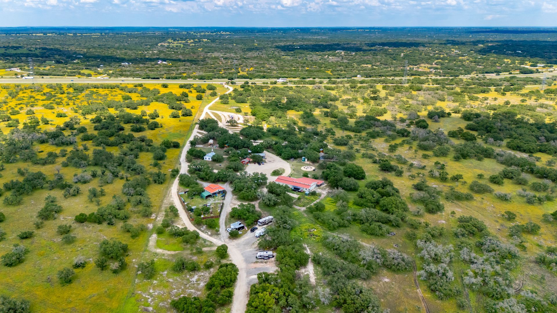 Aerial view of a rural landscape with dirt roads, scattered green shrubs, yellow wildflowers, and a small house.