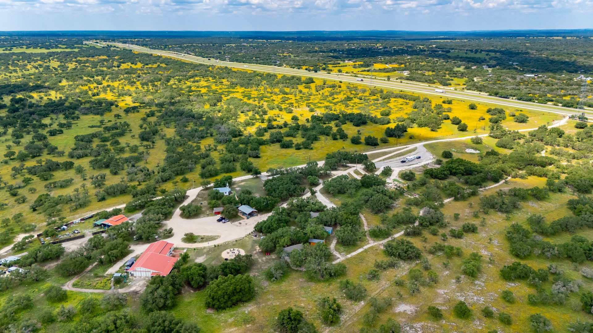 Aerial view of a rural landscape with green fields, yellow wildflowers, scattered trees, and a few buildings.