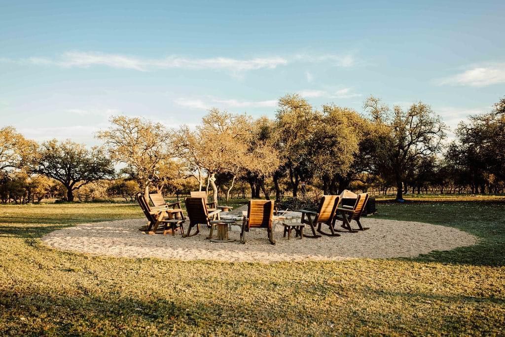 Circle of wooden chairs around a fire pit on a grassy field with trees in the background