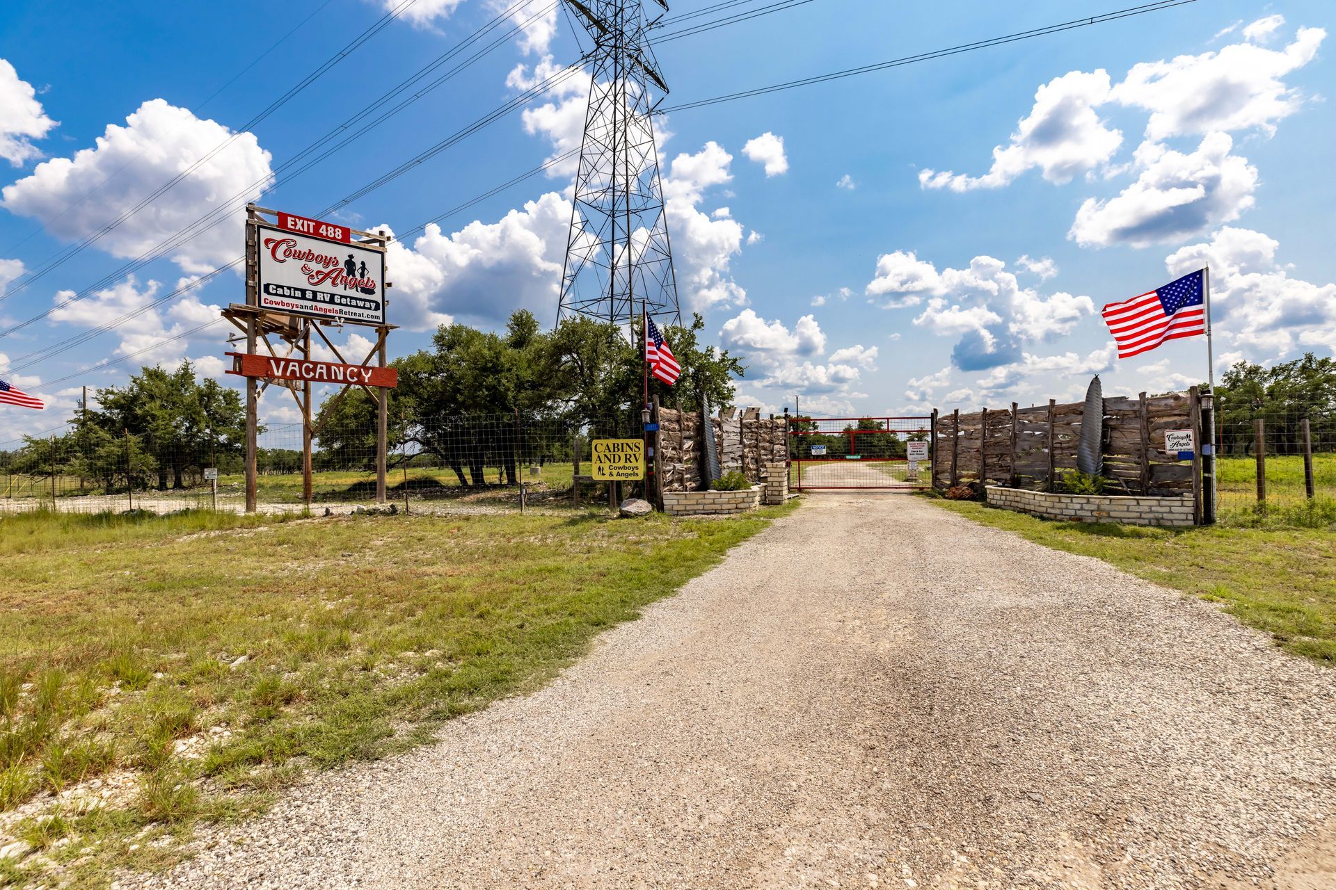Gravel road leading to a rustic ranch entrance with wooden fences and an American flag under a blue sky