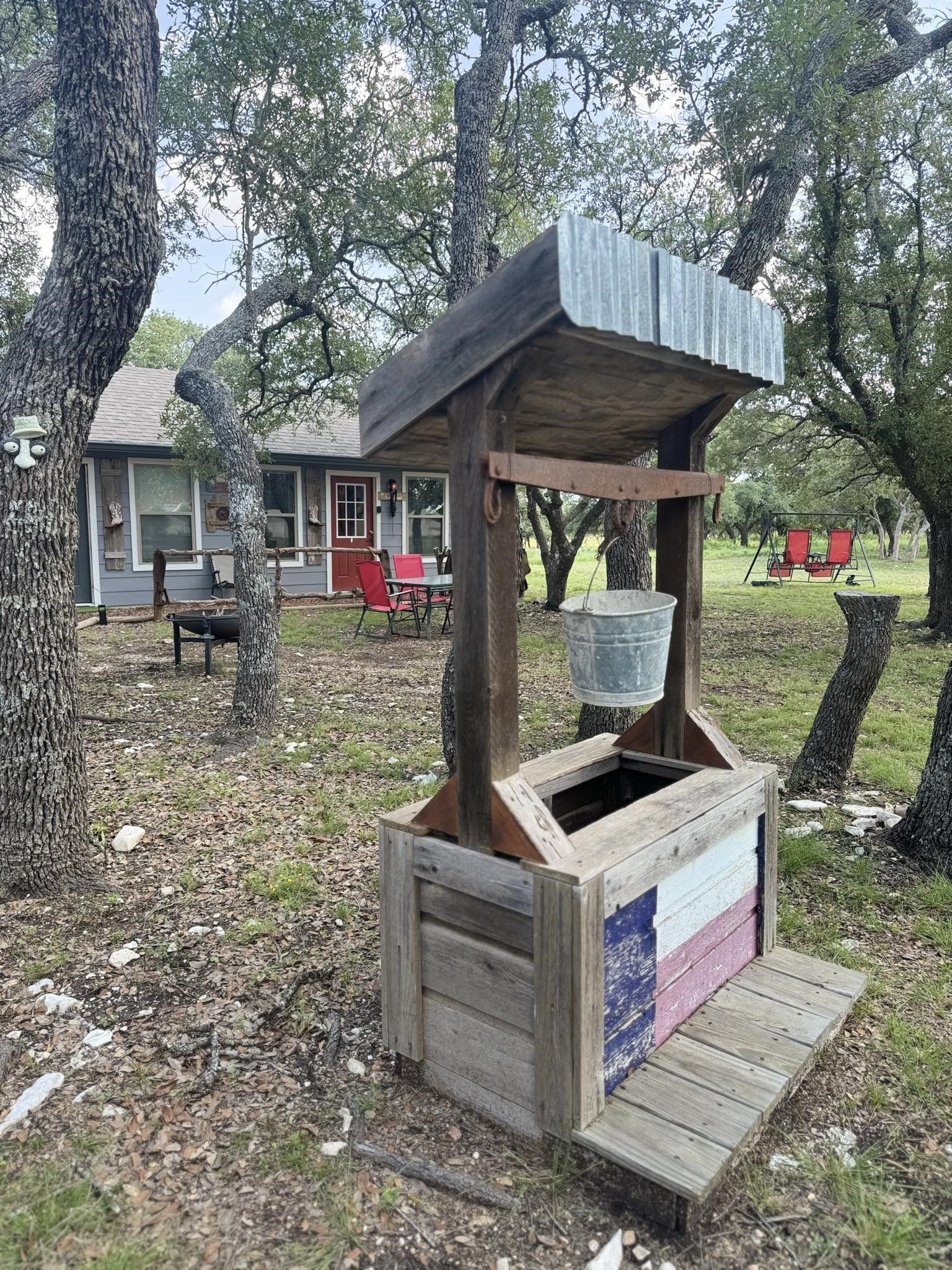 Rustic well with wooden roof and bench in a wooded yard beside a small cabin