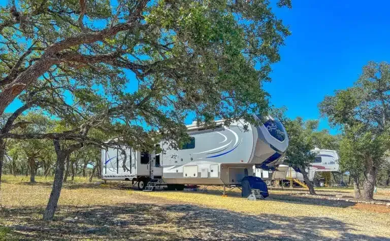 White RV trailer parked under trees in a sunny wooded area with a blue sky.
