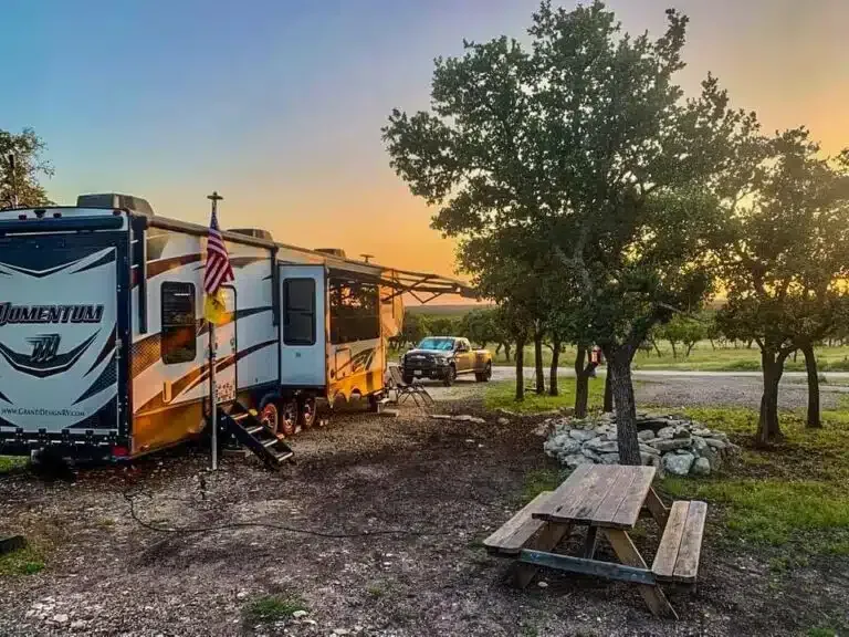 RVs parked at sunset near a picnic table and trees, with an American flag in a campground.