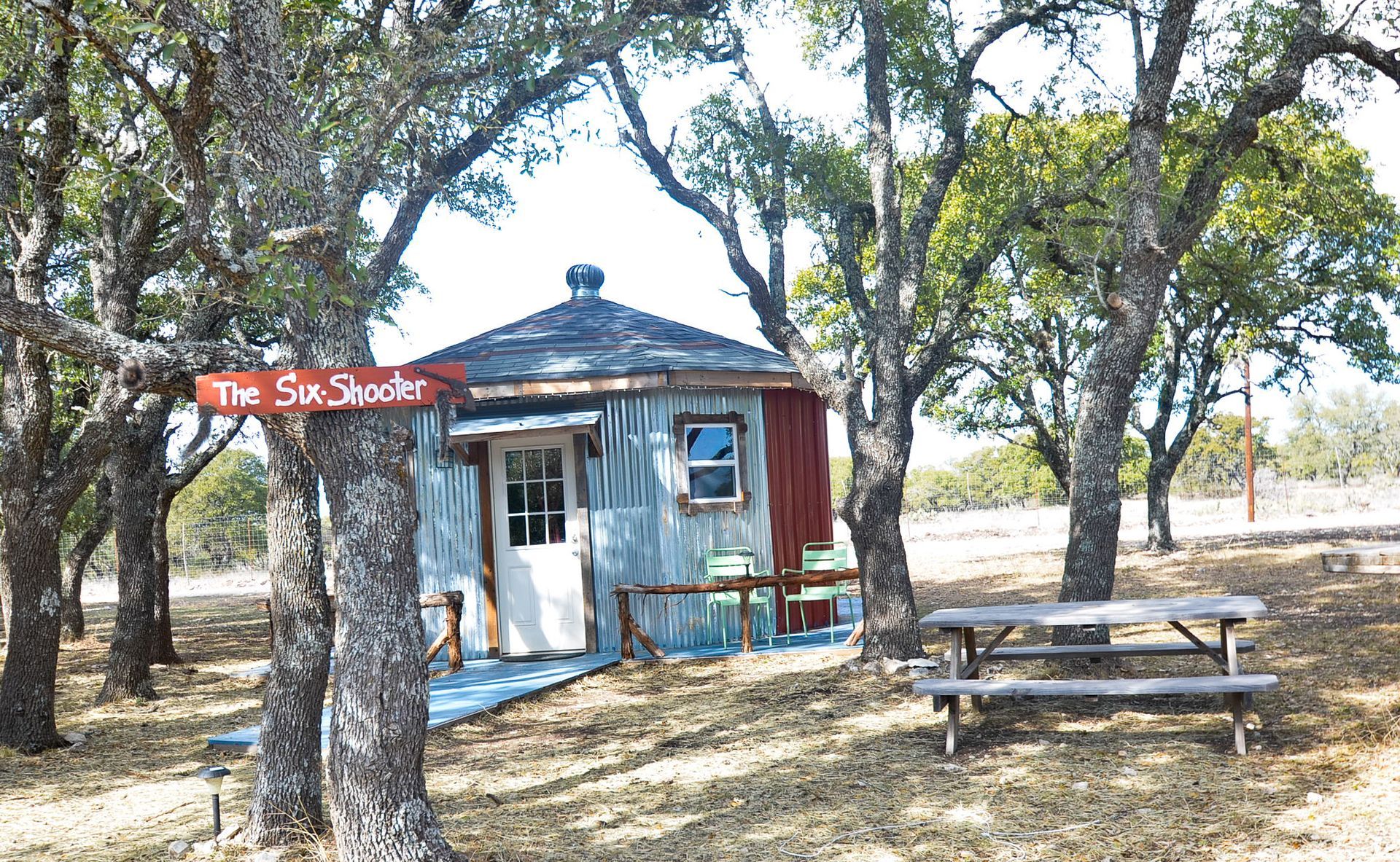 Small blue-and-red roadside shack with a sign among trees, beside a picnic table in a sunny clearing