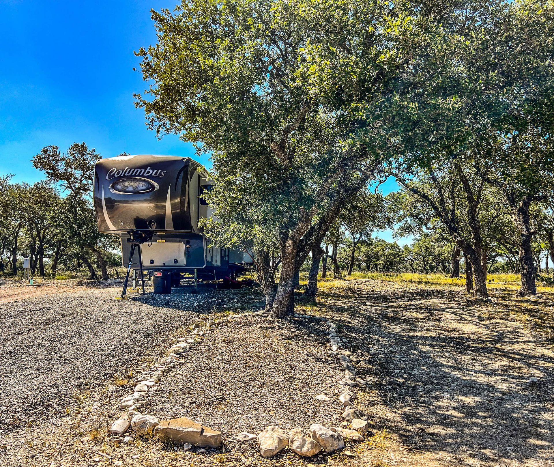 Parked RV under leafy trees in a sunny gravel campsite