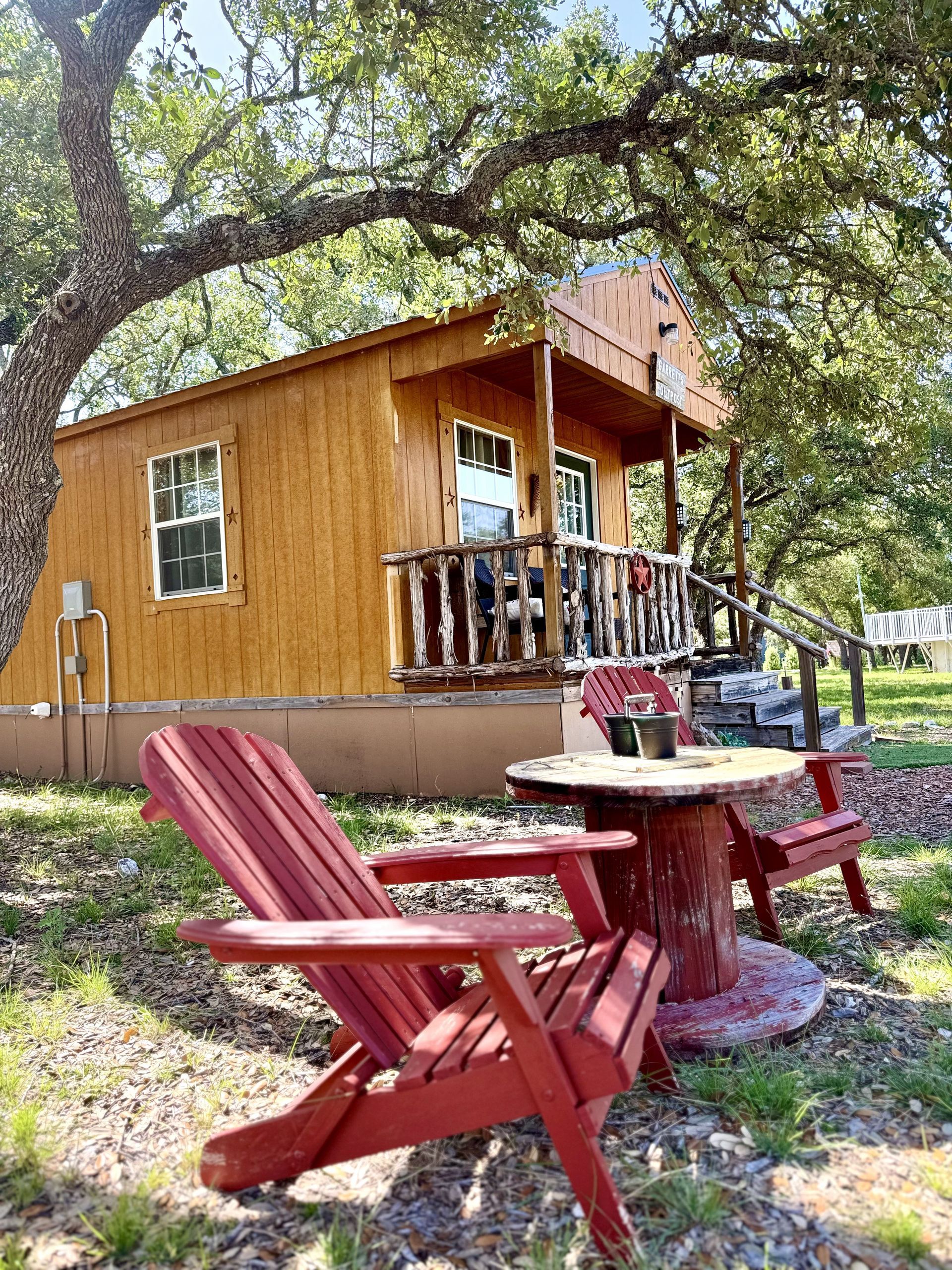 Red Adirondack chairs around a small table beside a wooden cabin under a large tree.
