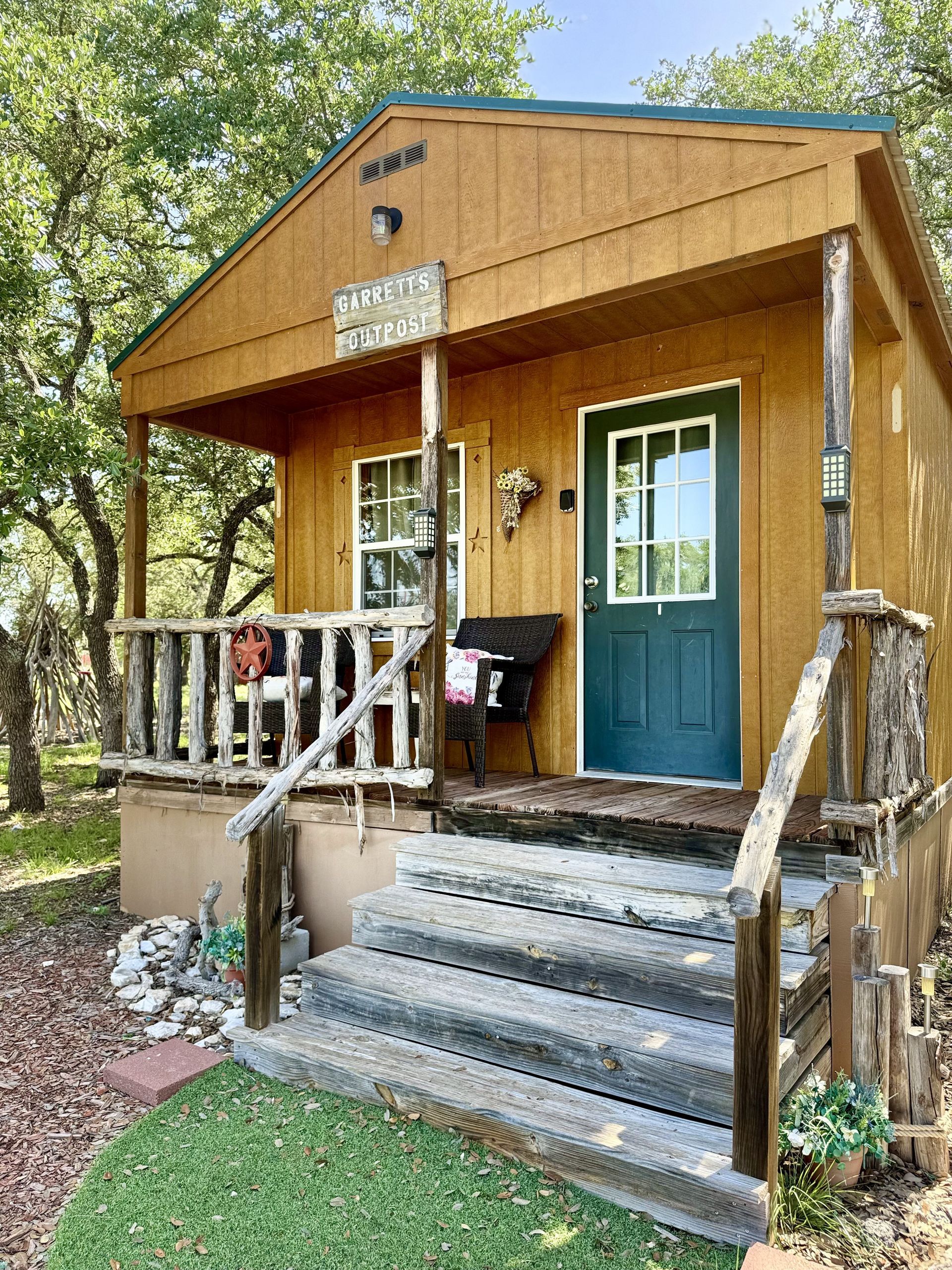 Small rustic wooden cabin with a blue door and front steps, surrounded by trees and grass.