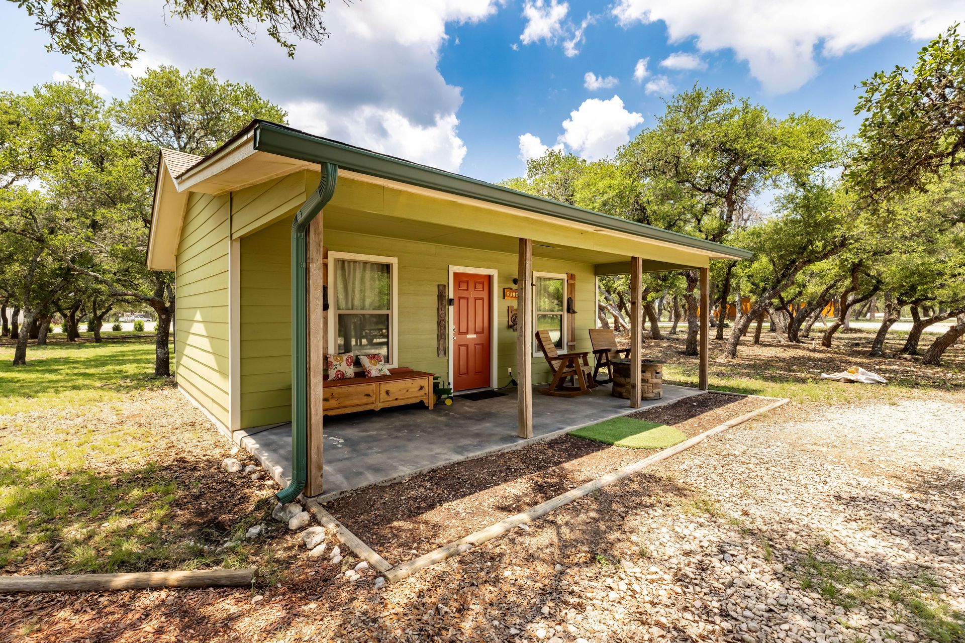 Small green cabin with a porch in a wooded yard under a bright blue sky