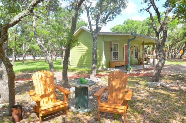 Two wooden chairs beside a small table under trees, in front of a green house with a porch