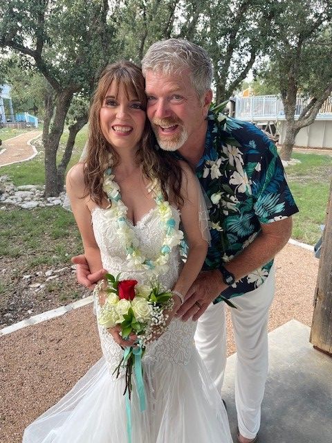 Couple posing outdoors at a wedding, smiling and holding a floral bouquet.