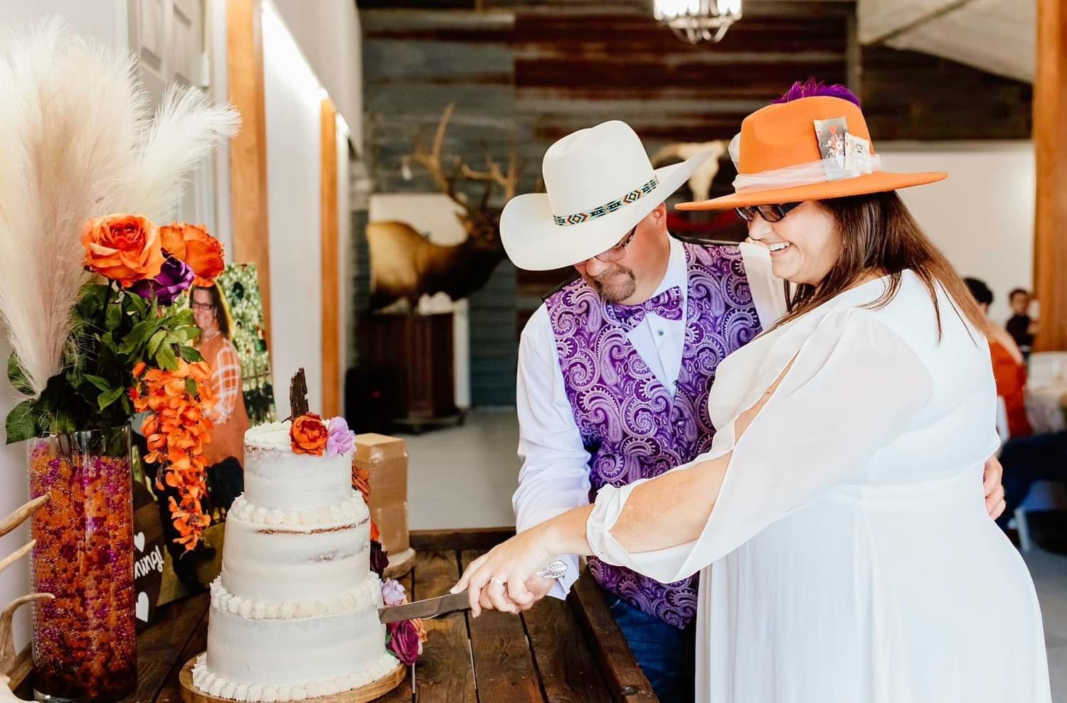 Couple in western hats cutting a white wedding cake beside orange floral decorations