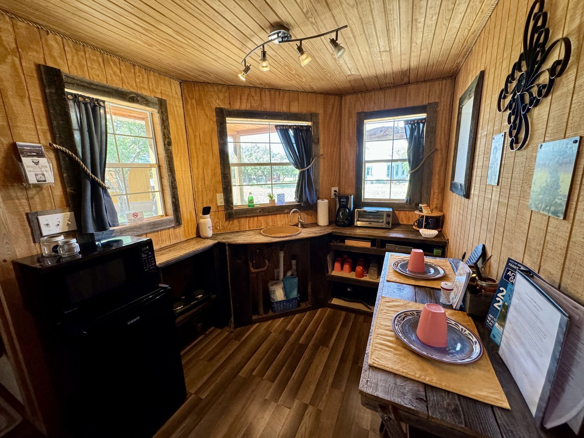 Cozy wood-paneled kitchen with black cabinets, three windows, and a small table with dishes