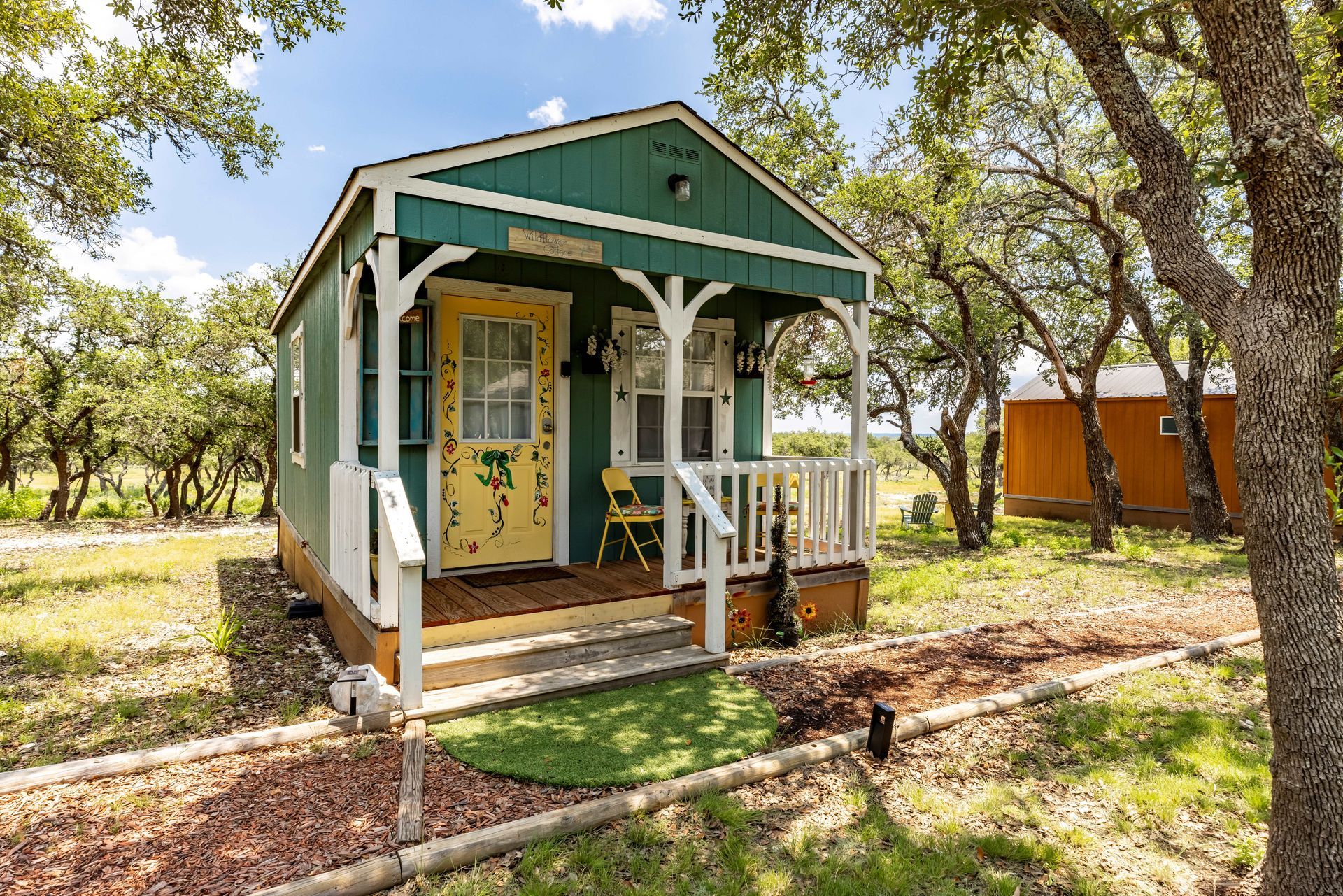 Small teal cabin with yellow door on a wooded path, with white porch railings and a green ramp.