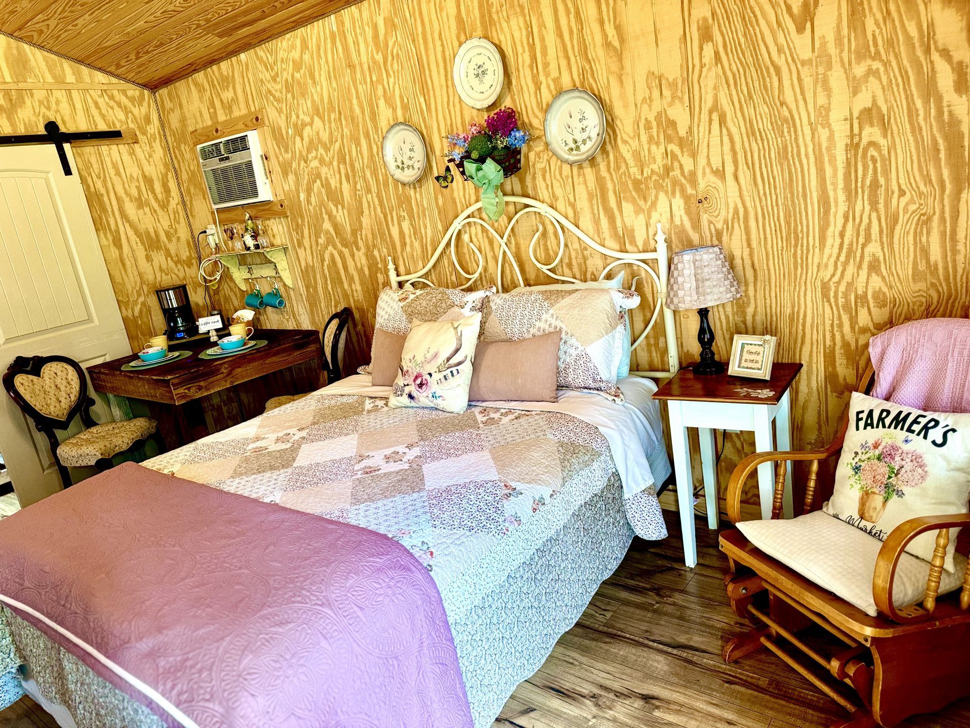 Cozy bedroom with white iron bed, lavender quilt, wicker chair, and wood-paneled walls