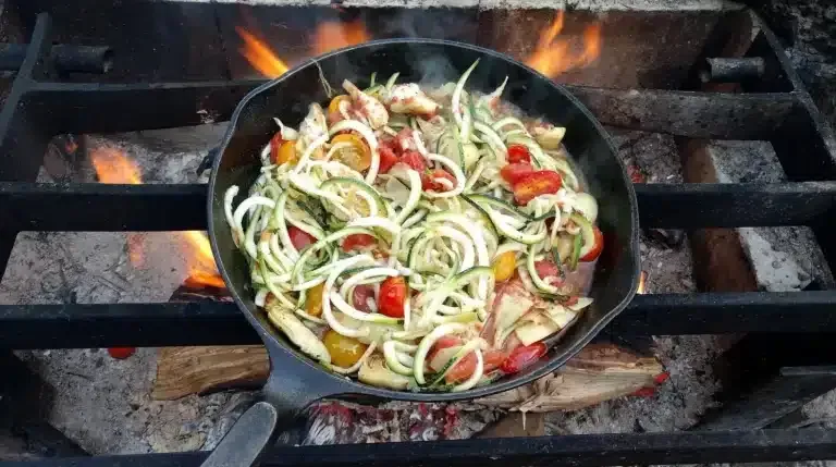 Skillet of sliced vegetables sizzling over a campfire on a grill, with flames visible beneath.