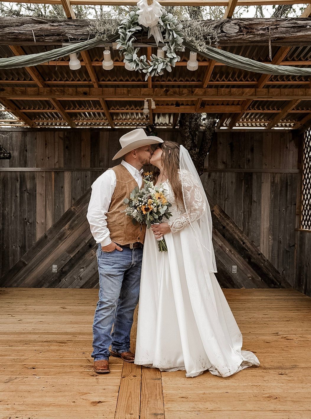 Wedding couple kissing on a rustic wooden stage, bride in flowing white dress holding bouquet, groom in cowboy hat and boots