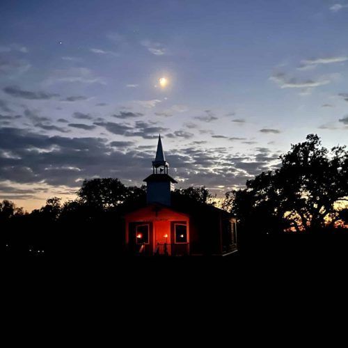 Small church at dusk with glowing red windows beneath a moonlit, cloudy sky