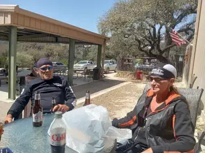 Two people sit at an outdoor picnic table with drinks under a shaded patio.