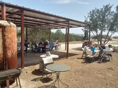 Outdoor shaded seating area with people at tables beside a dirt clearing and trees