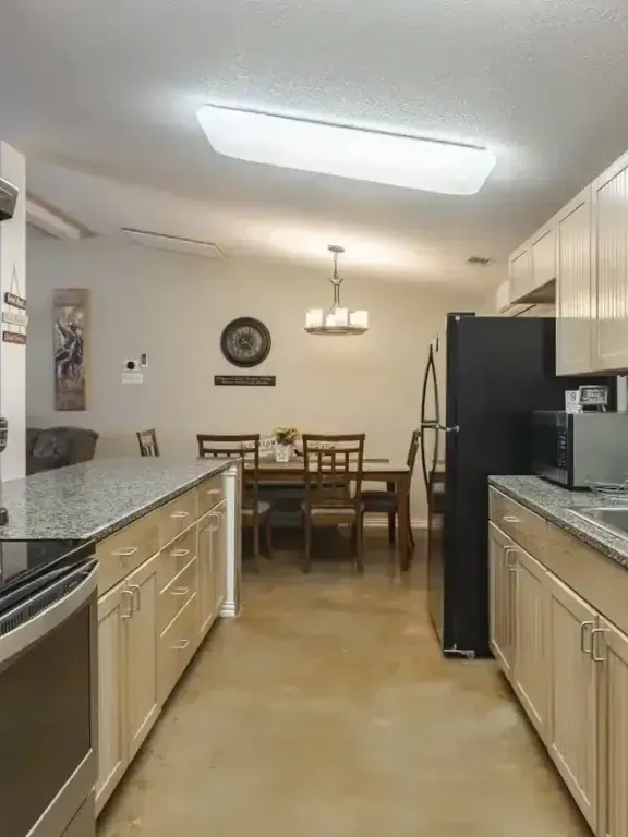 Long galley kitchen with beige cabinets, granite counters, black appliances, and a dining table at the far end