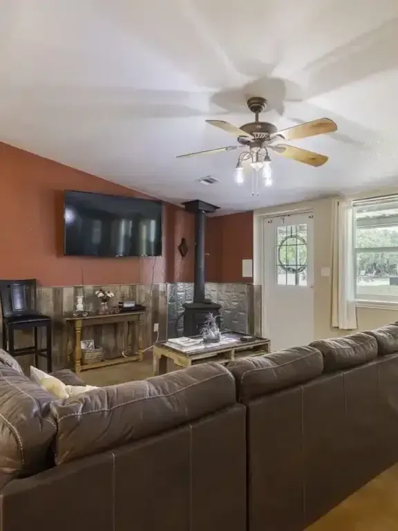 Living room with a large brown sectional, wall-mounted TV, ceiling fan, and stone fireplace near a bright window.