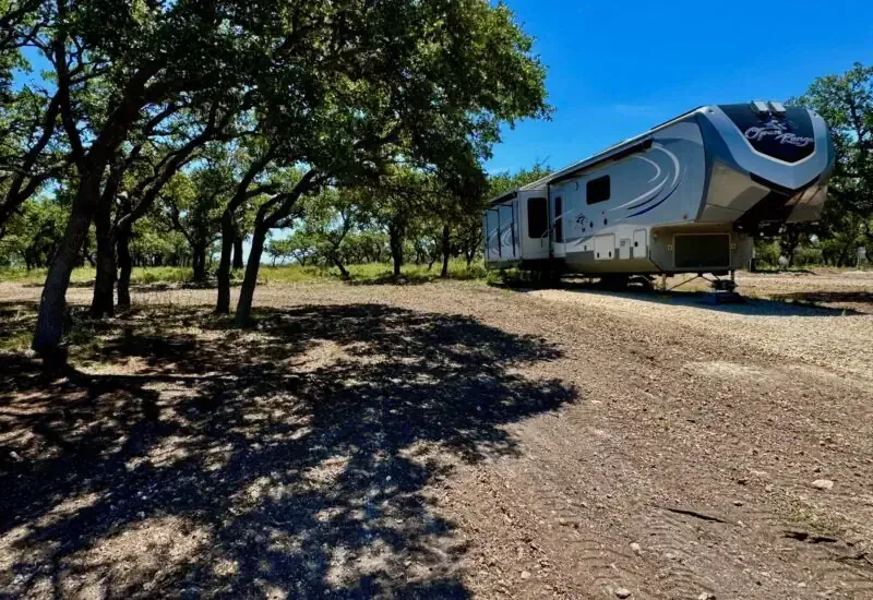 RV parked on a gravel campsite under large trees in bright sunlight