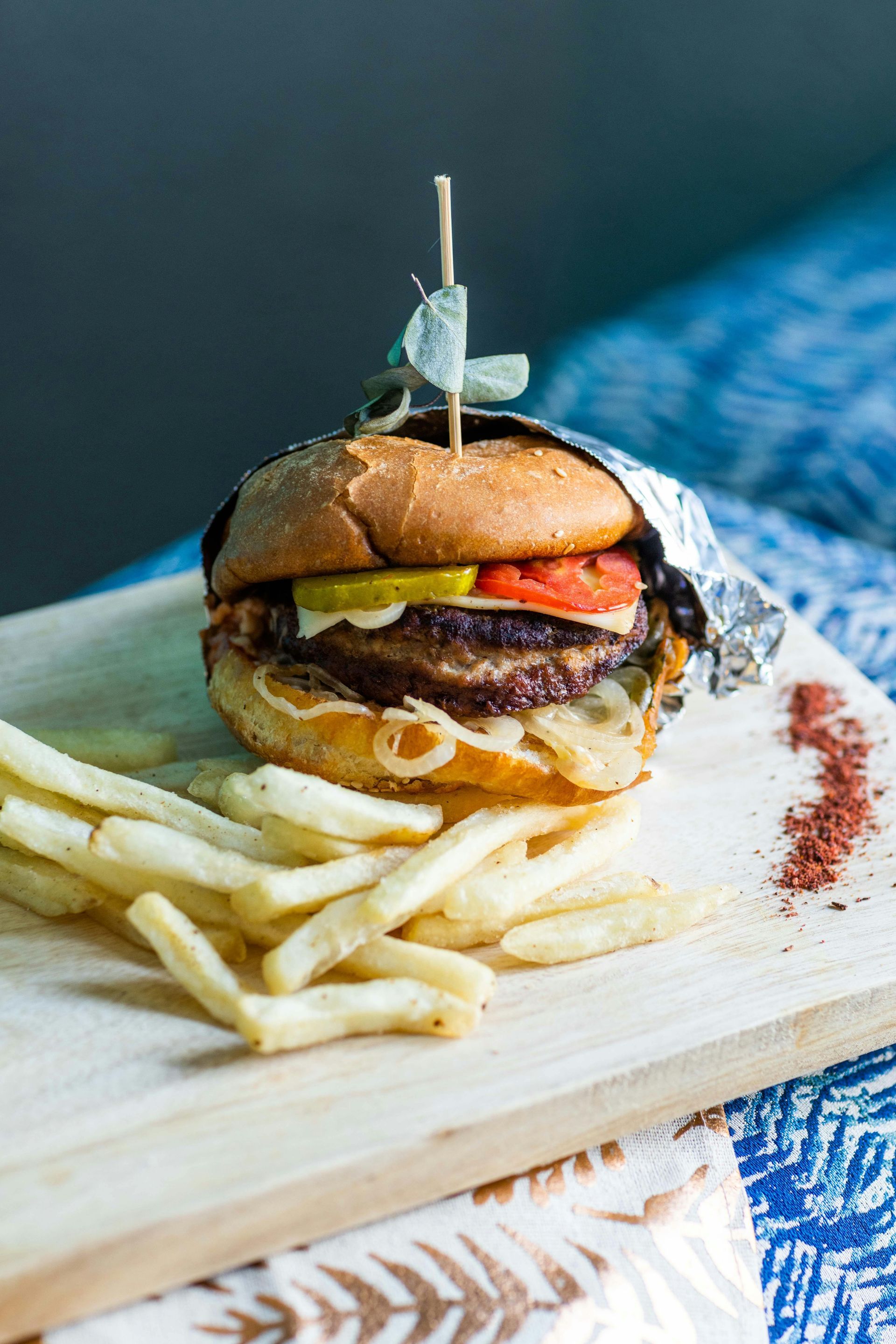 Burger with fries on a wooden board, topped with a skewer, on a blue patterned cloth.
