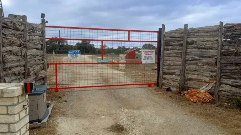 Red metal gate between stacked stone walls blocking a gravel road under a cloudy sky