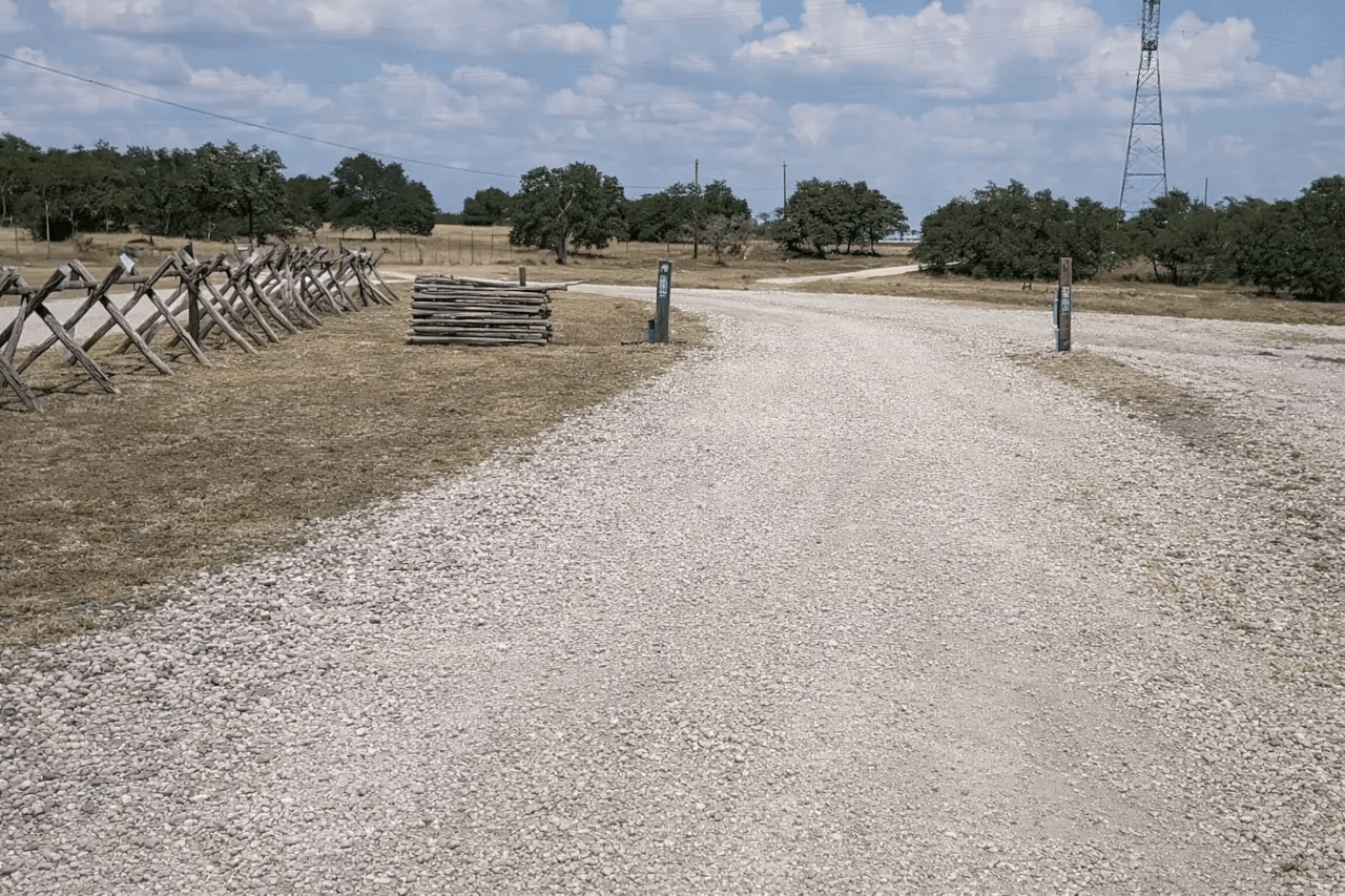 Gravel road through a dry field with scattered trees and a small barricade on the left