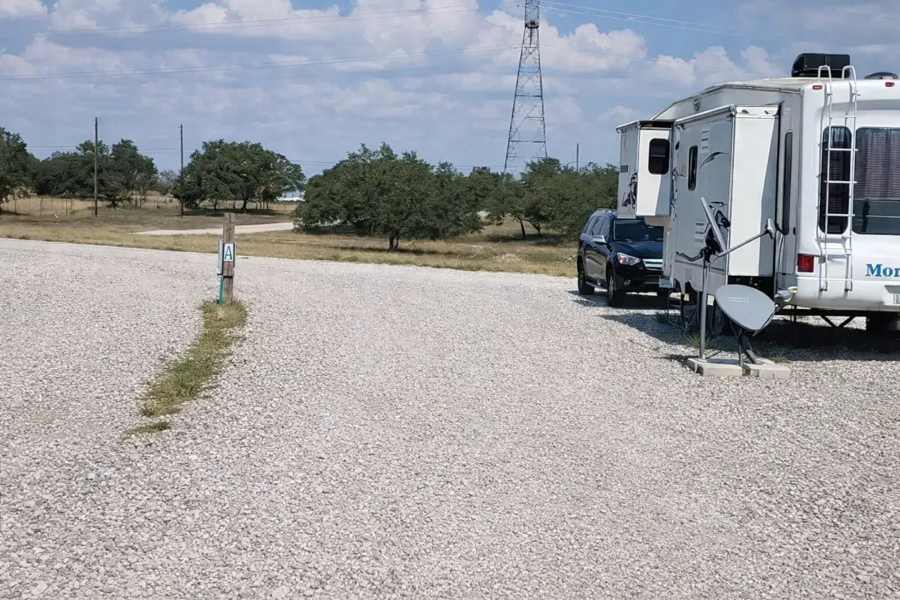 White RV parked on a gravel lot beside a blue truck, with trees and a power tower in the background